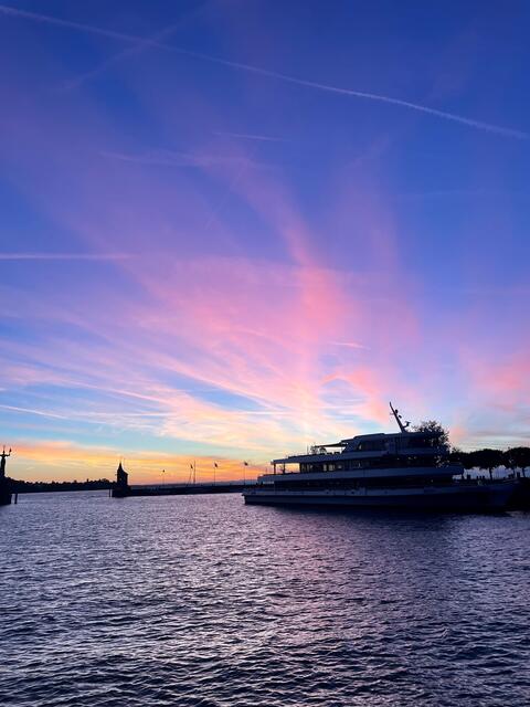 Blick in den Konstanzer Hafen an einem Morgen im Oktober mit einem Farbenspiel am Himmel in blau, pink und gelb. Im Hafen liegt ein Schiff.