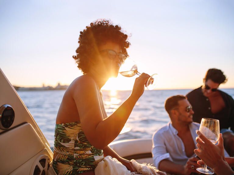 Frau mit einem Glas Wein auf Deck von einem Schiff der BSB. Im Hintergrund sind zwei Männer und der Bodensee.