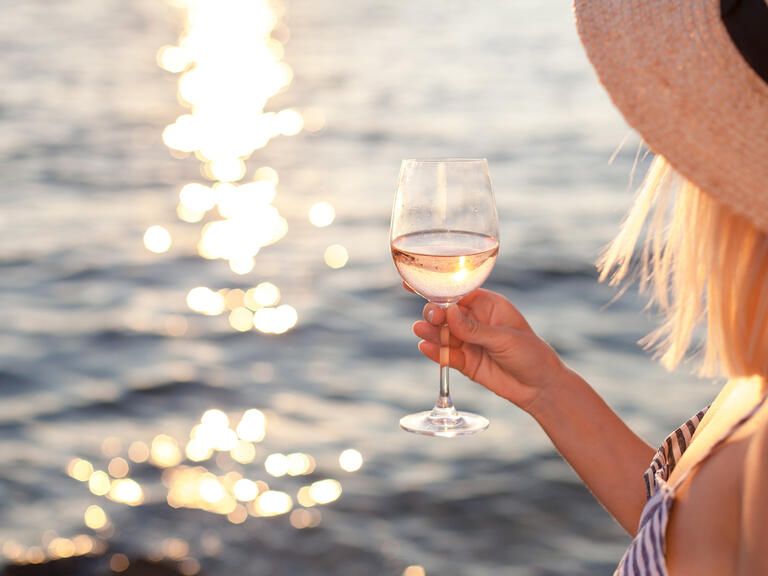 Frau hält Weinglas mit Weisswein in der Hand. Im Hintergrund die Sonne über Wasser.