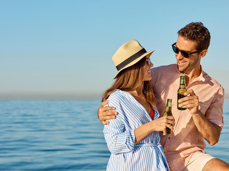 Eine Frau und ein Mann stoßen Arm in Arm mit einem Bier an. Im Hintergrund ist Wasser, die Sonne scheint, beide tragen Sonnenbrillen.
