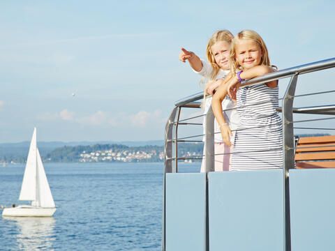 Zwei Kinder an Bord einer Fähre, im Hintergrund das blaue Wasser des Bodensees und ein Segelboot.